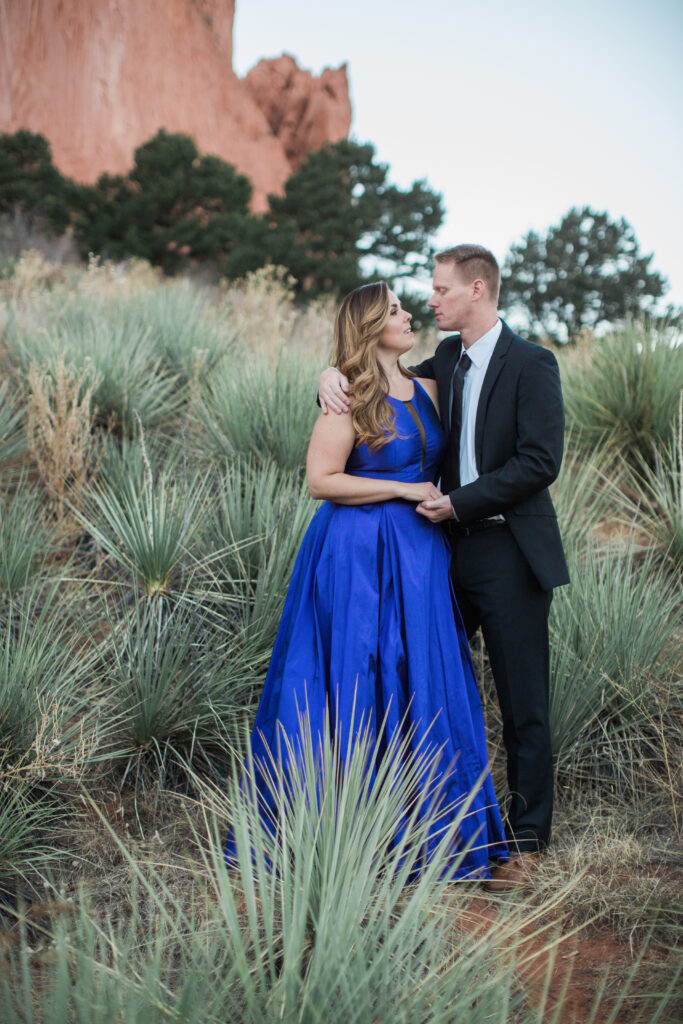 couple wearing blue fancy dress and black suit standing in embrace near red rocks 