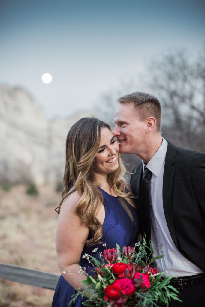 couple wearing blue fancy dress and black suit standing in embrace near red rocks with moon in background