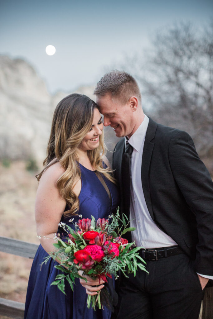 couple wearing blue fancy dress and black suit standing in embrace near red rocks with moon in background