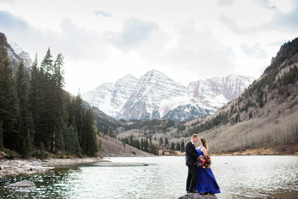 couple wearing blue fancy dress and black suit standing in embrace at lake with mountains in the distance 