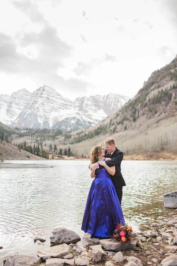 couple wearing blue fancy dress and black suit standing in embrace at lake with mountains in the distance 