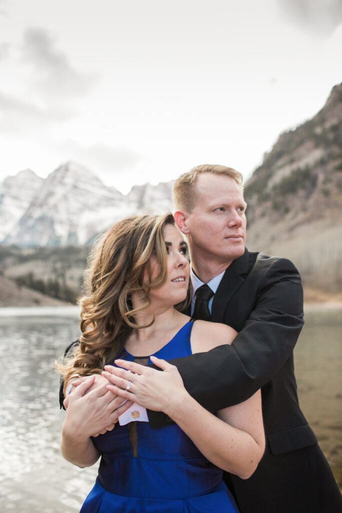 couple wearing blue fancy dress and black suit standing in embrace at lake with mountains in the distance 