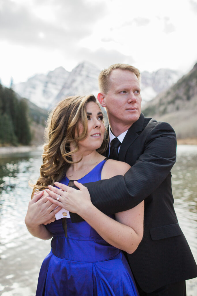 couple looking off into distance wearing blue fancy dress and black suit standing in embrace at lake with mountains in the distance 