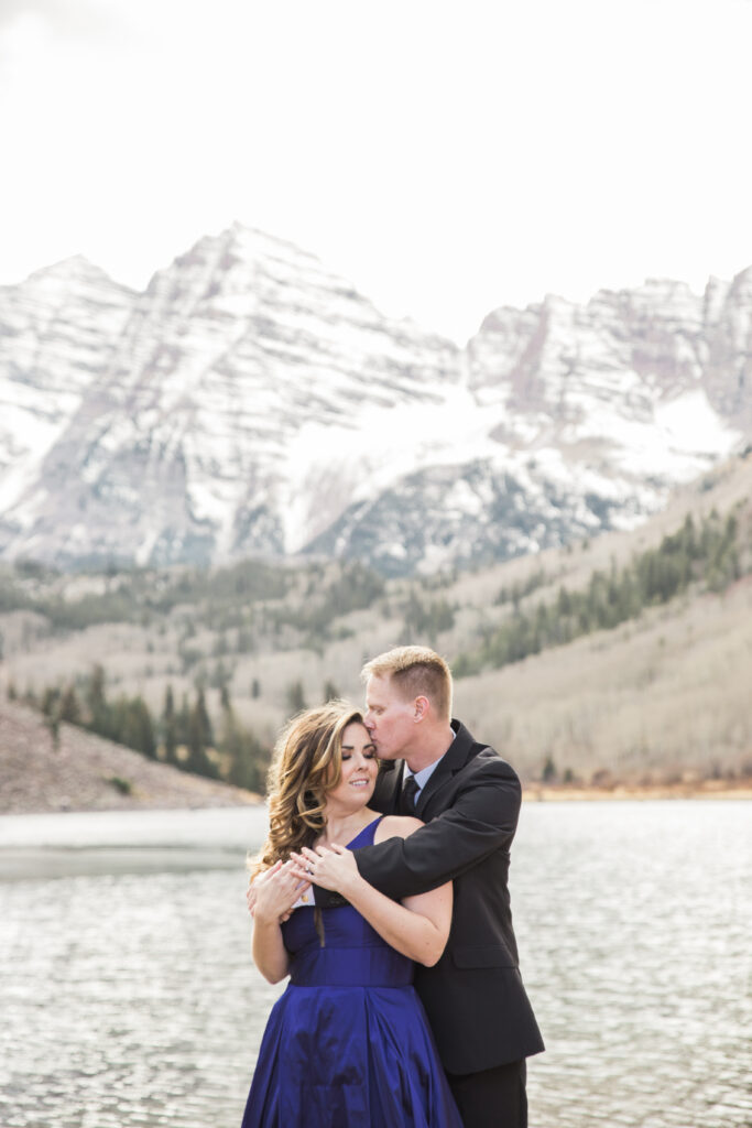 couple wearing blue fancy dress and black suit standing in embrace at lake with mountains in the distance 