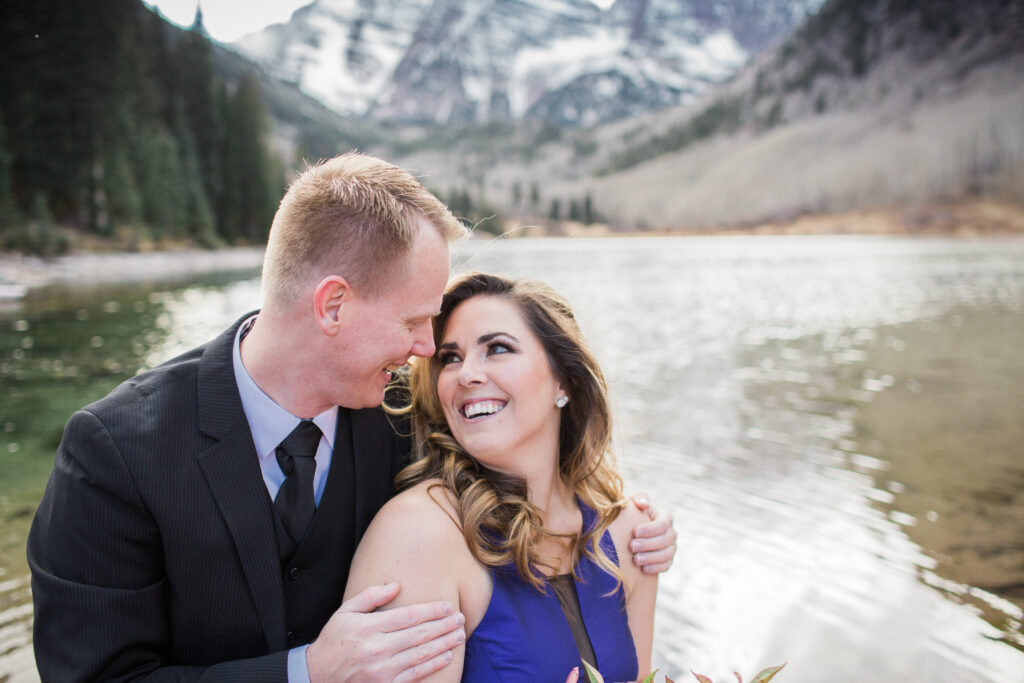 couple wearing blue fancy dress and black suit standing in embrace at lake with mountains in the distance 