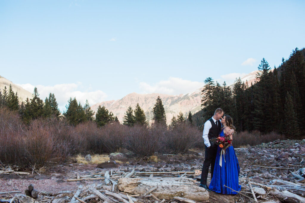 couple wearing blue fancy dress and black suit standing in embrace at lake with mountains in the distance 