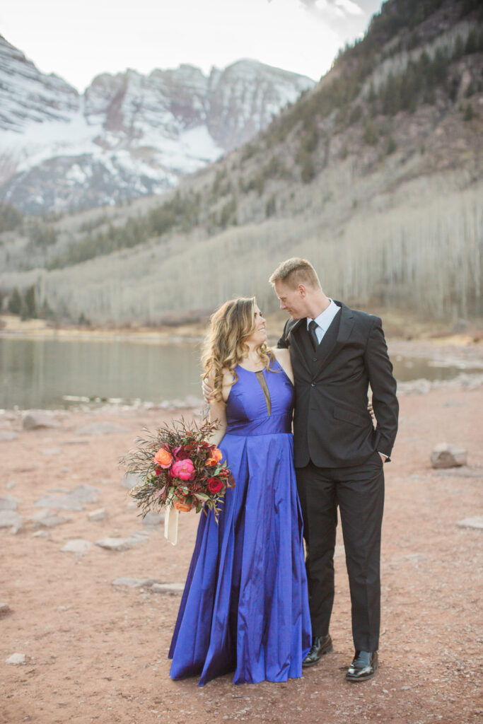 couple wearing blue fancy dress and black suit standing in embrace at lake with mountains in the distance 