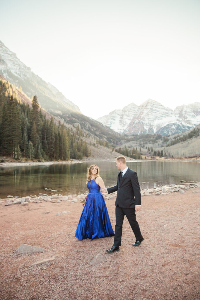 couple wearing blue fancy dress and black suit standing in embrace at lake with mountains in the distance 