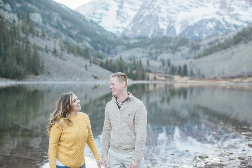 couple laughing and holding hands by the lake with mountains in the background 