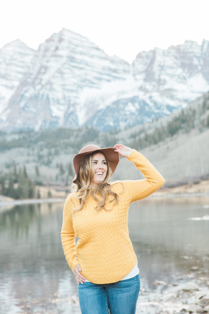 woman smiling wearing yellow sweater and a floppy brown hat with mountains in the distance 