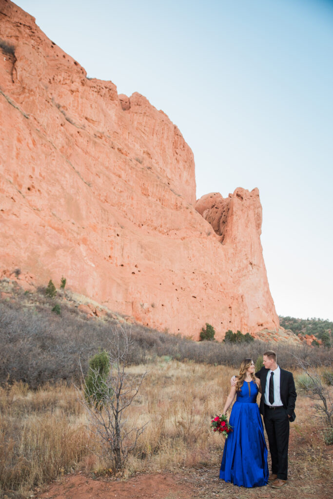 couple wearing blue fancy dress and black suit standing in embrace near red rocks 