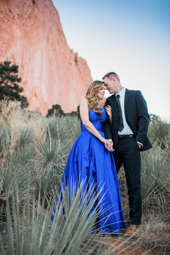 couple wearing blue fancy dress and black suit standing in embrace near red rocks 