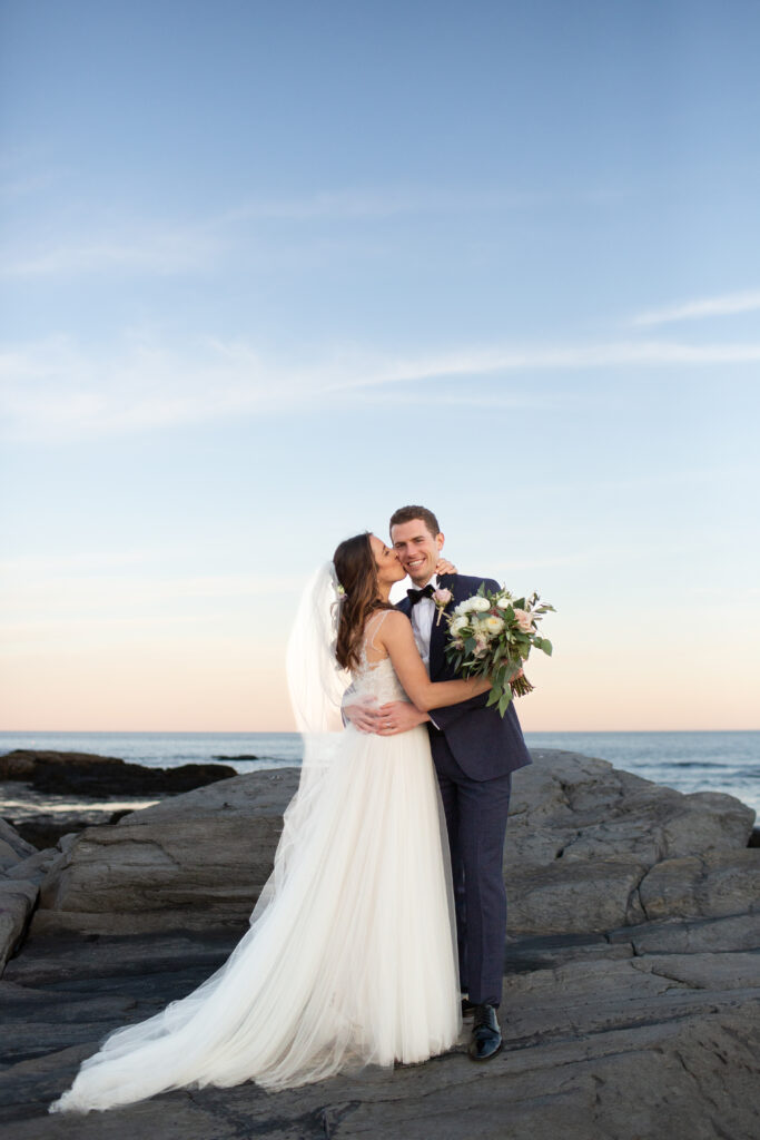 bride kissing grooms cheek while they stand on the rocks with the ocean in the background