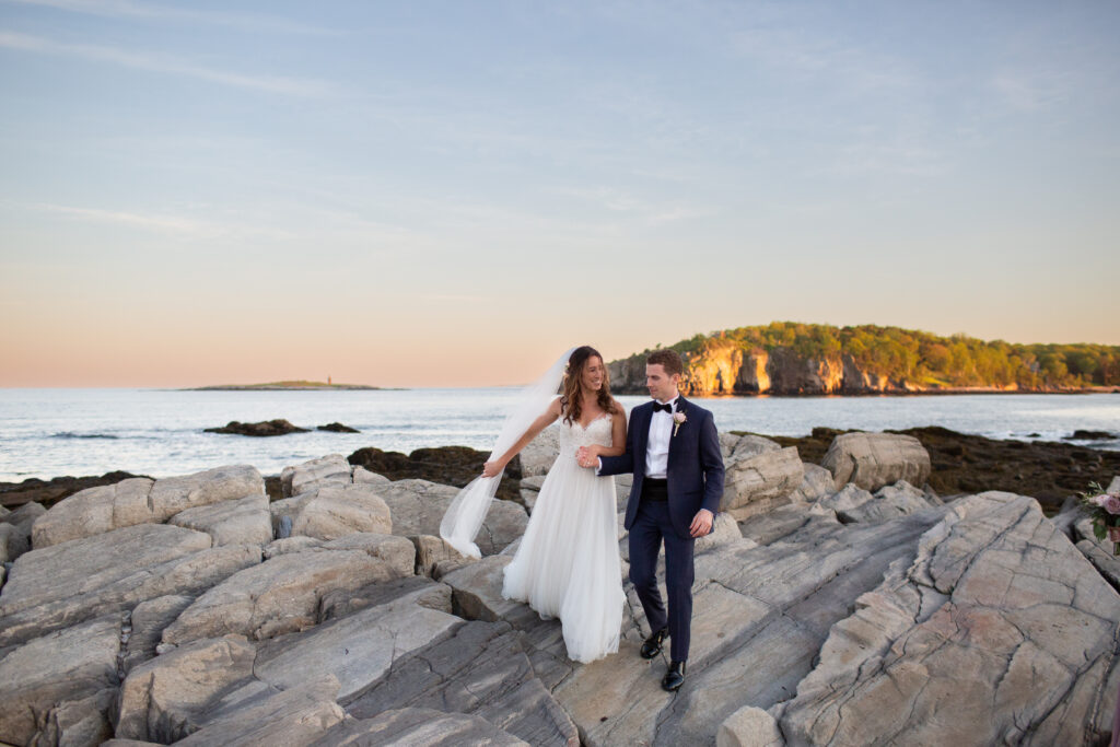 bride grabbing veil as it blows in the wind while they walk off of the rocks at the water 