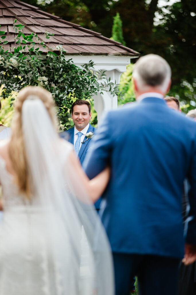 bride being walked down the aisle with her dad, groom seen through them photo from the back