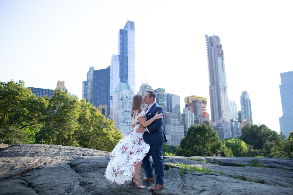 couple kissing in central park with skyline in the background wind is blowing her dress