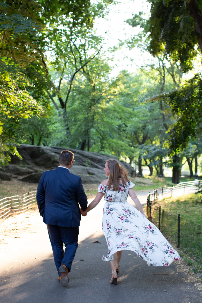 couple walking holding hands in central park and the wind is blowing her dress behind her 