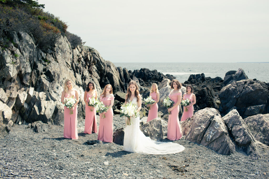 bridesmaids in pink dresses with bride standing on rocky maine beach 