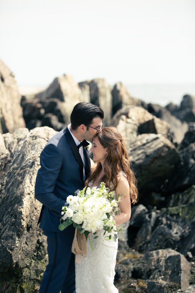 bride and groom standing on rocky Maine beach with groom kissing brides forehead 