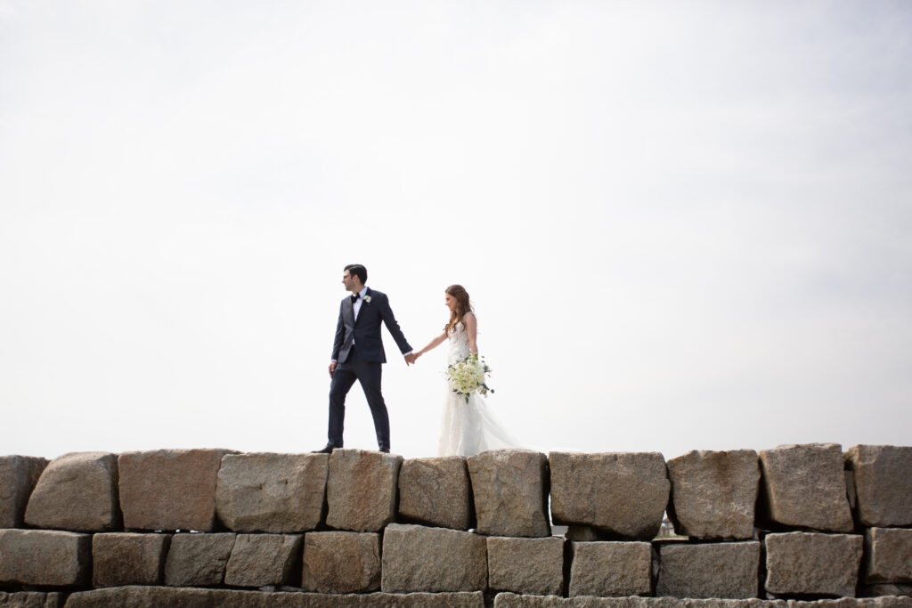 bride and groom walking on rocky jetty holding hands 